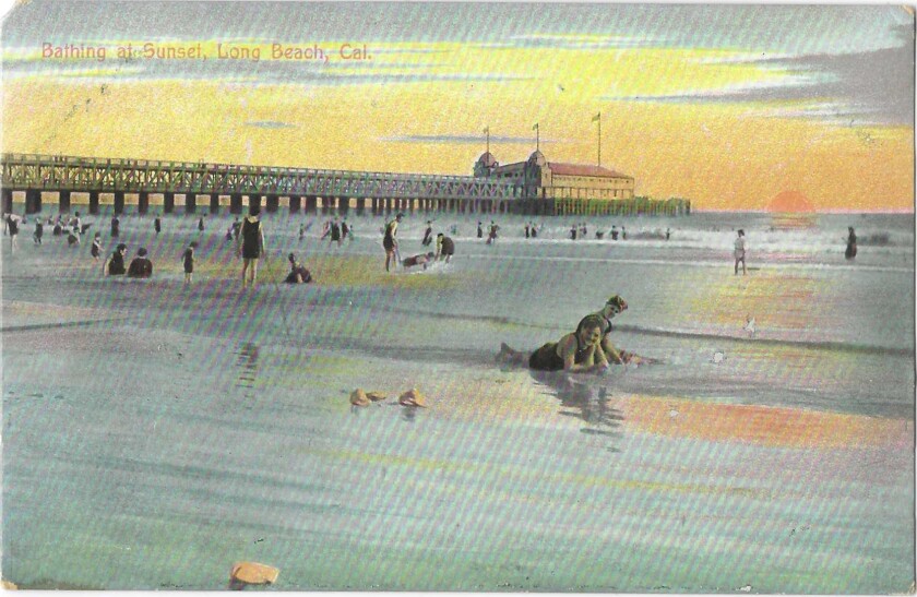 In the foreground, two people lie in the wet sand as the sun sets on other bathers and a pier behind them.