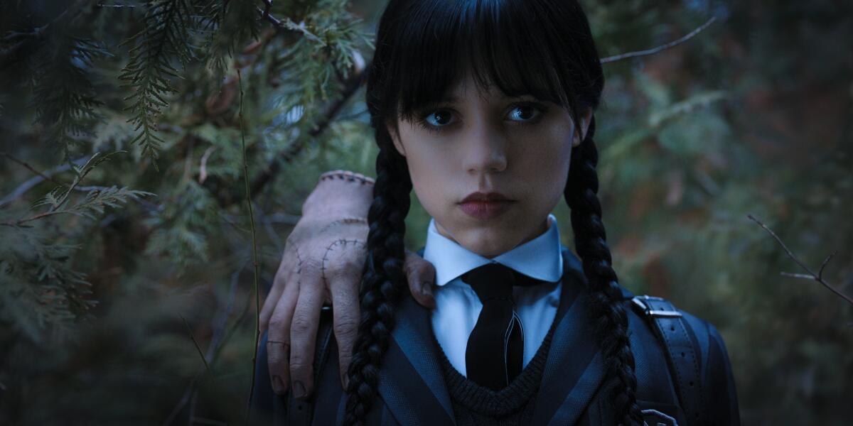 A young woman with long, dark pigtail braids has a floating hand perched on her shoulder in “Wednesday.”