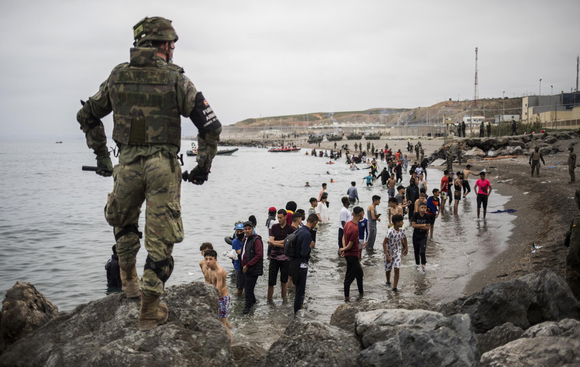 A military member in camo fatigues carrying a baton stands on a rock overlooking dozens of migrants exiting the sea