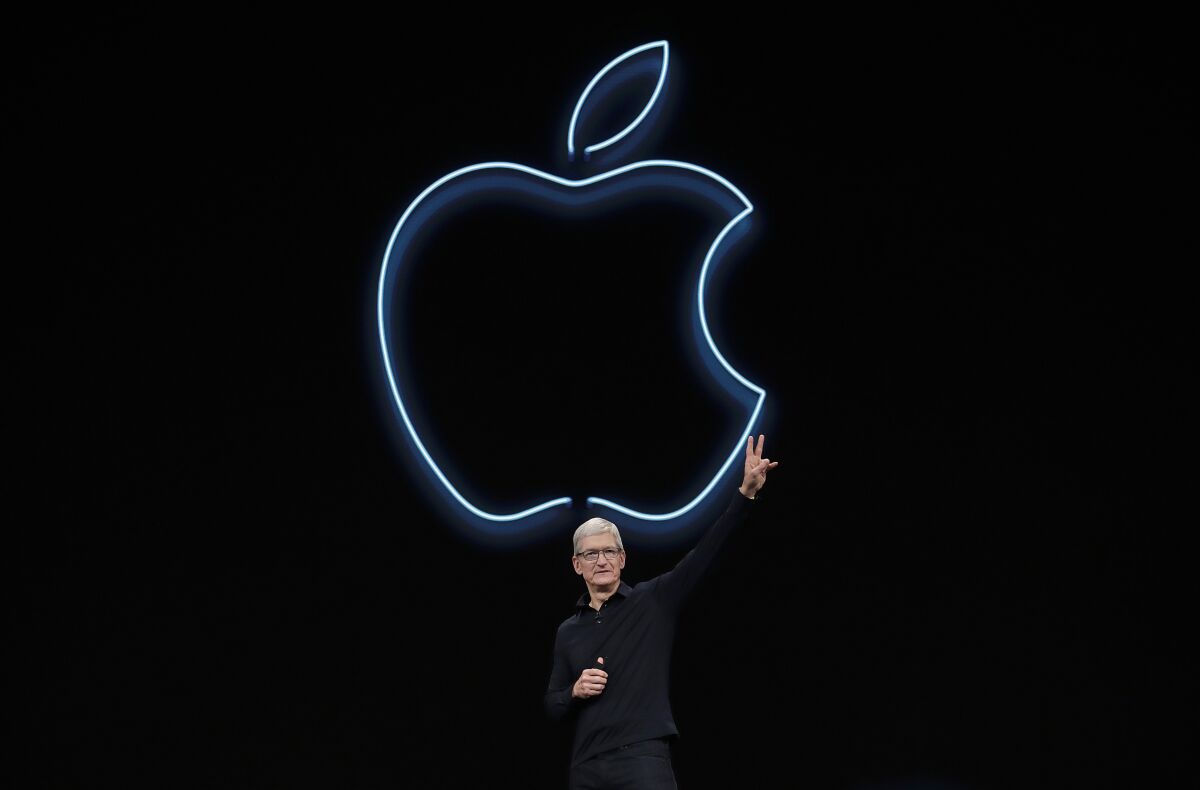 A man dressed in black and standing in front of an illuminated outline of an apple gives a peace sign 