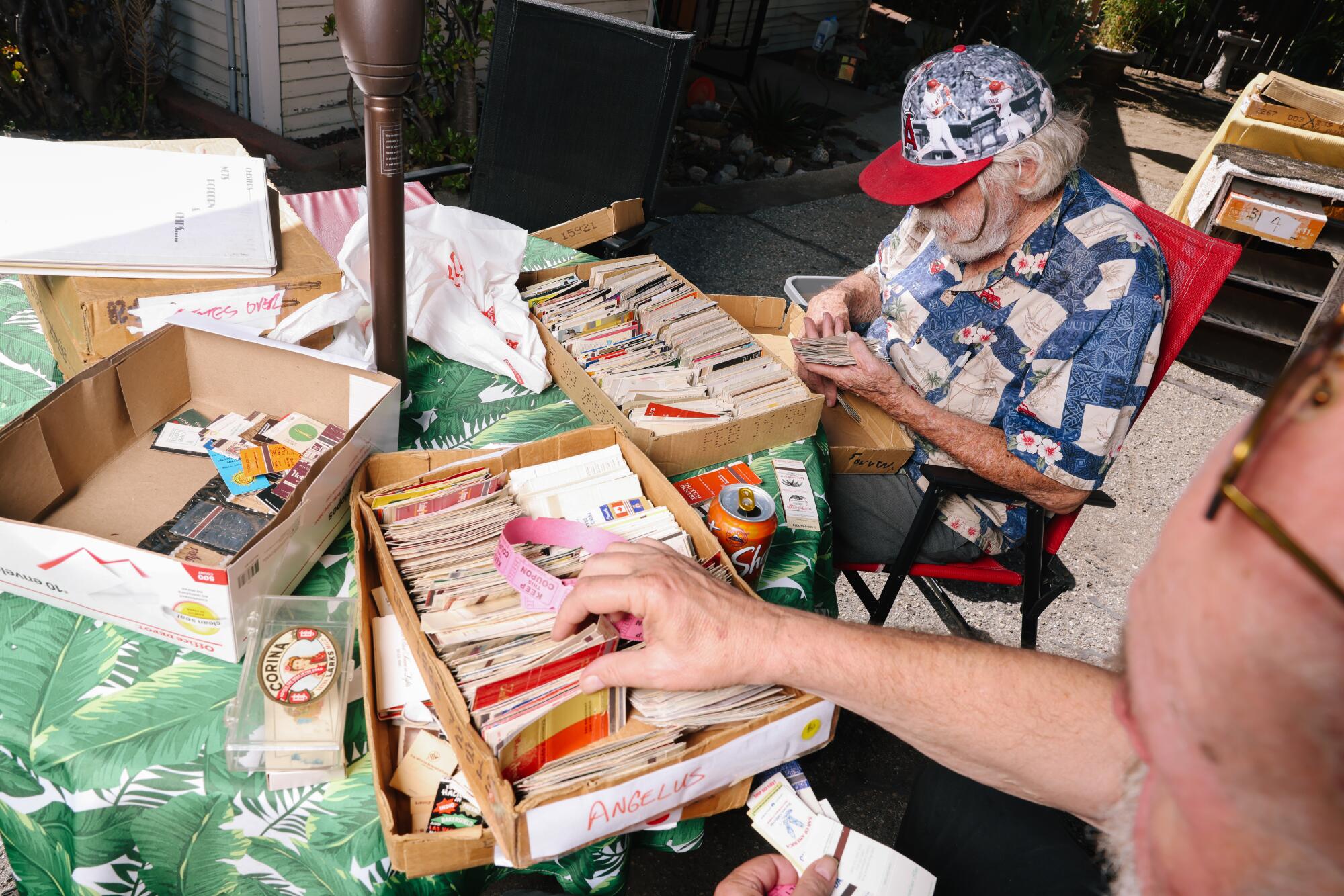 Two men seated at a table sort through boxes of matchbook covers.