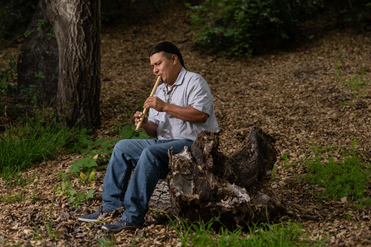 A man plays the flute in a wooded setting.