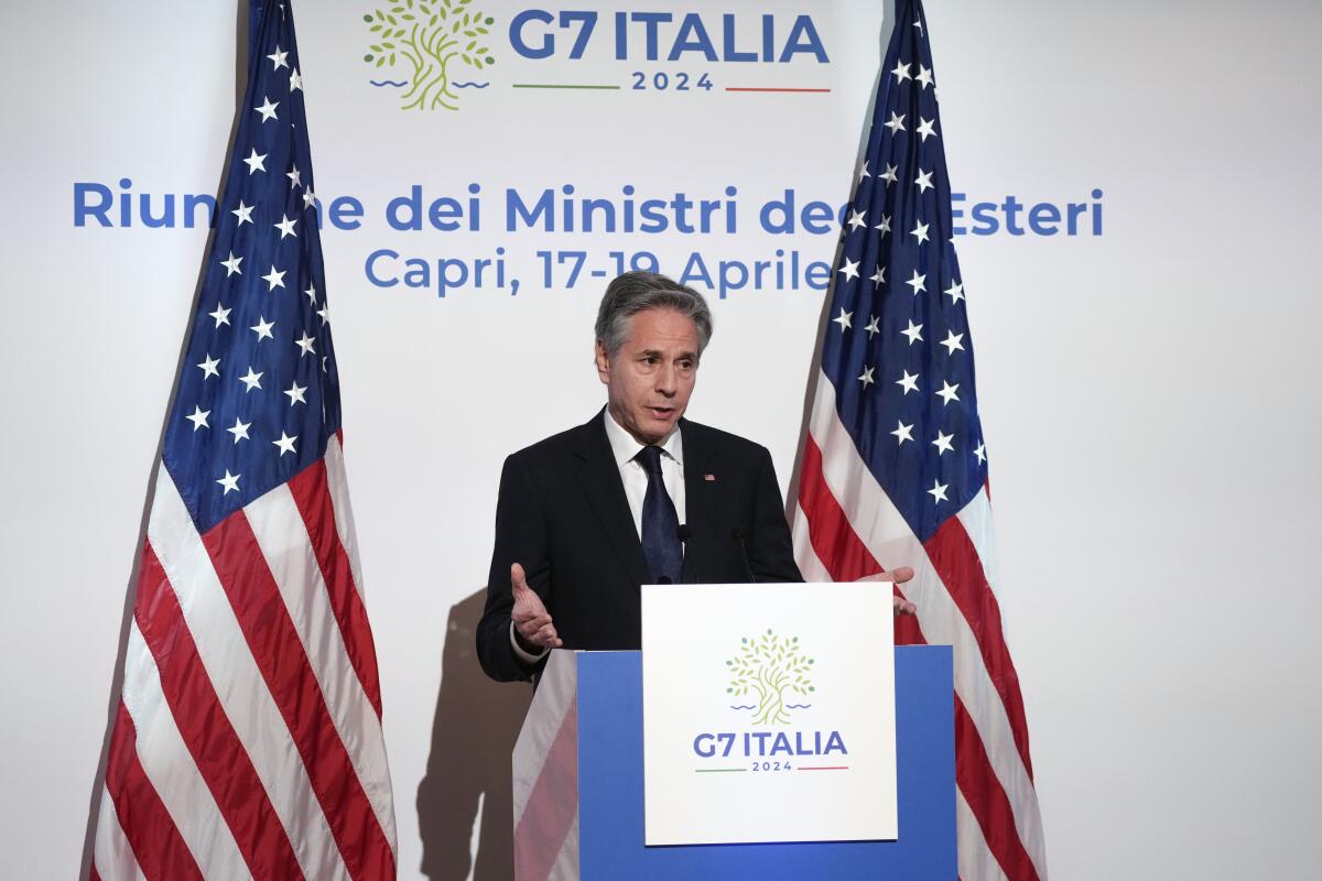 Secretary of State Antony J. Blinken stands at a dais between two American flags at a G7 news conference.