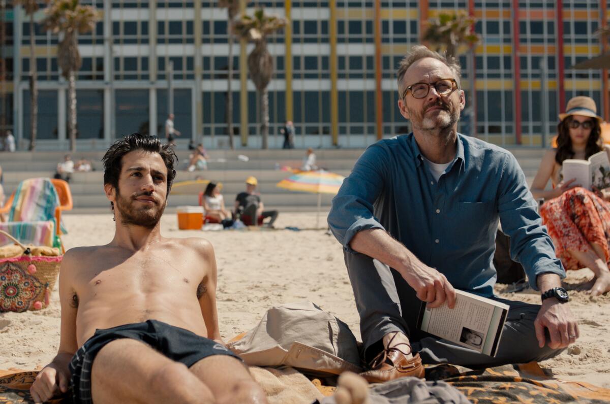 Niv Nissim, left, in swimsuit, and John Benjamin Hickey, in street clothes, sit on the beach