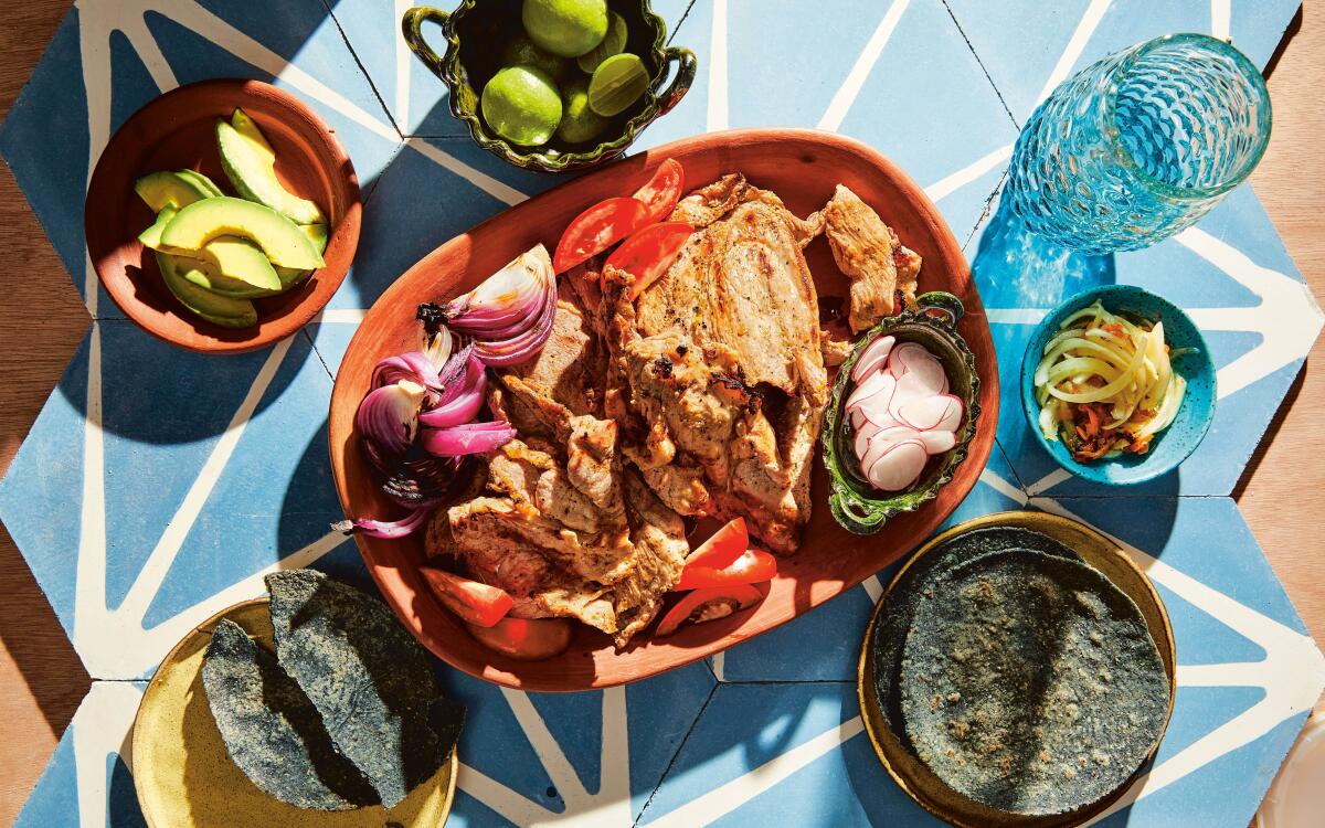 Thinly sliced pork shoulder surrounded by blue corn tortillas and cut avocado, limes, radishes and onions.