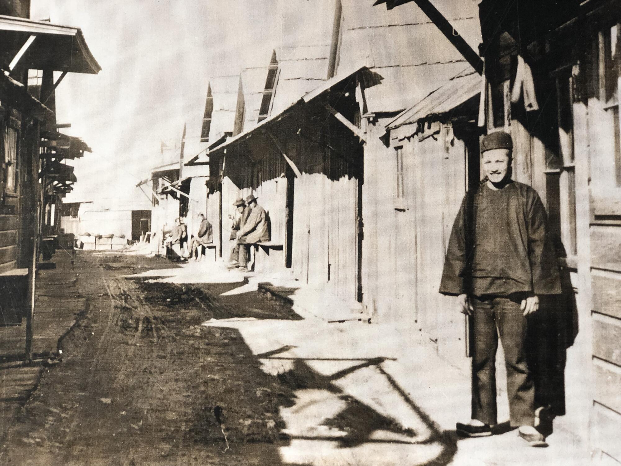 A black-and-white photo of a man in Chinese street clothes standing as others sit out front of a row of wooden houses.