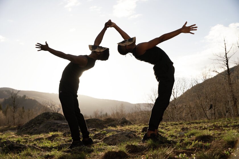 Two male dancers wearing decorative masks bend backward toward one another at the top of a hill. 