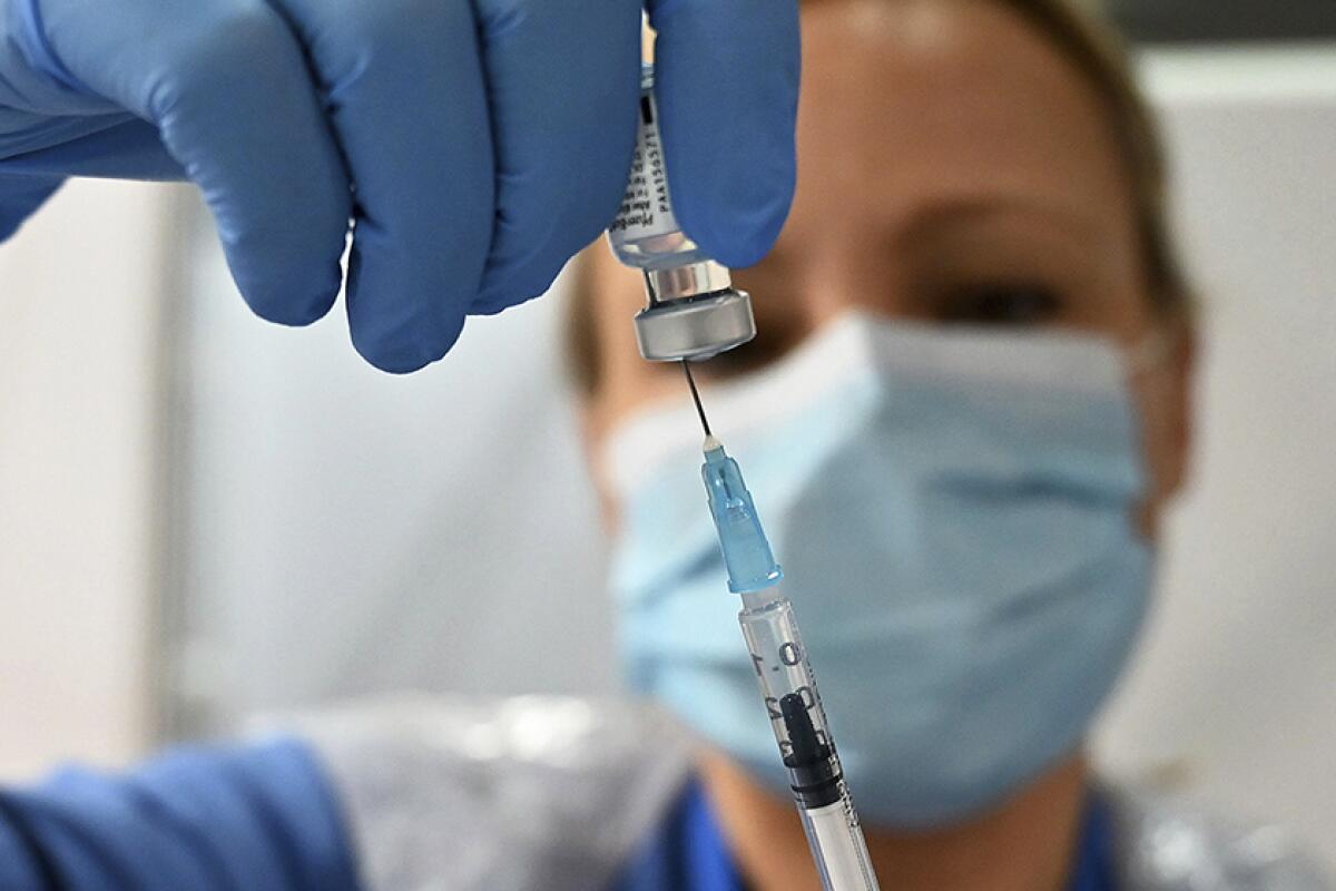 A masked woman wearing blue gloves inserts a needle into a vial of vaccine 
