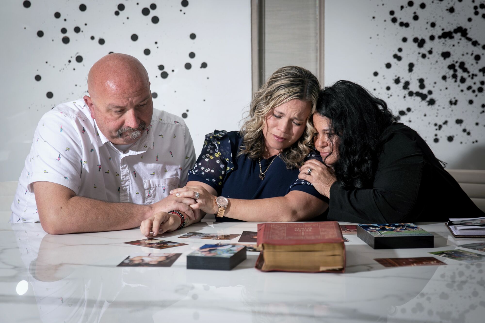 A man at left and two women, who are leaning on each other, sit behind a marble tabletop with photos laid out on it.