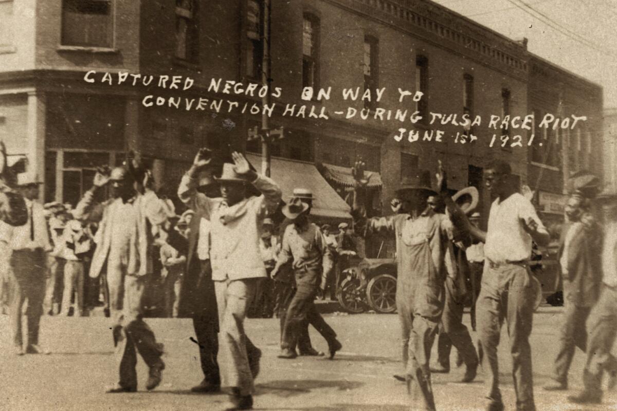 A group of Black men march down a street, hands in the air. Writing says they were "captured" in what was then called a riot.