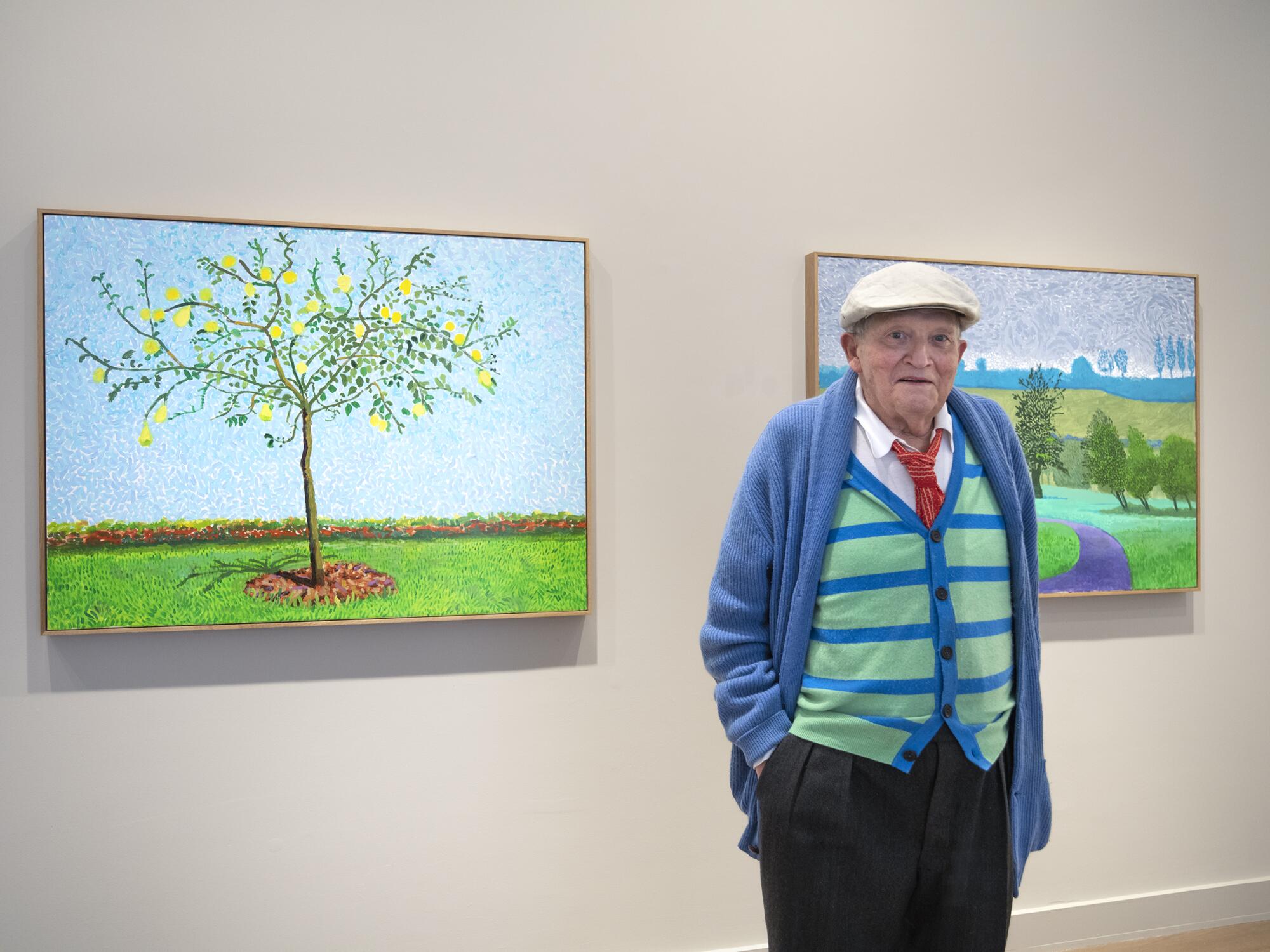 David Hockney, in striped sweater vest and cap, stands in front of two paintings hung on a gallery wall.