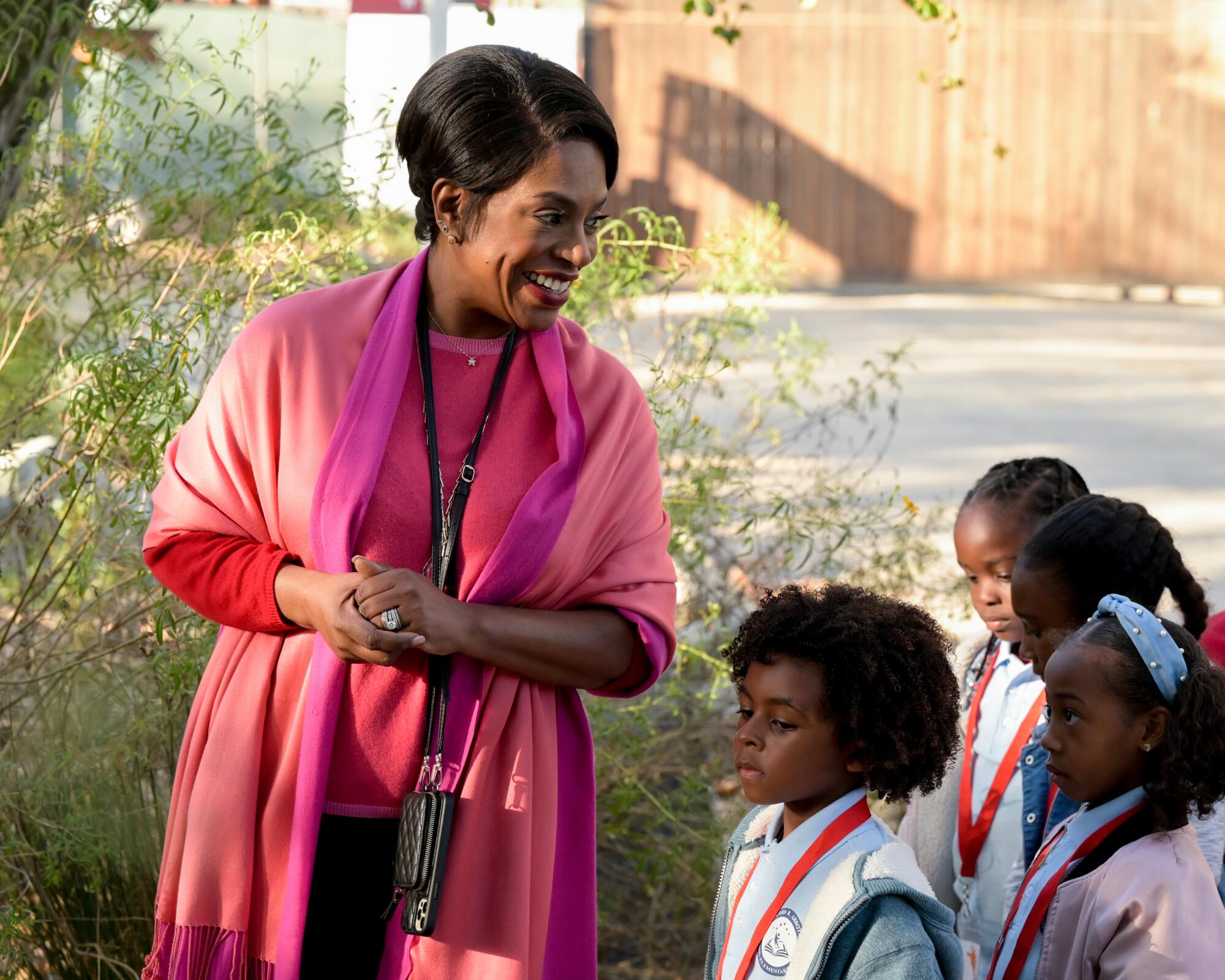 A woman dressed in pink stands leaning toward four young schoolchildren.