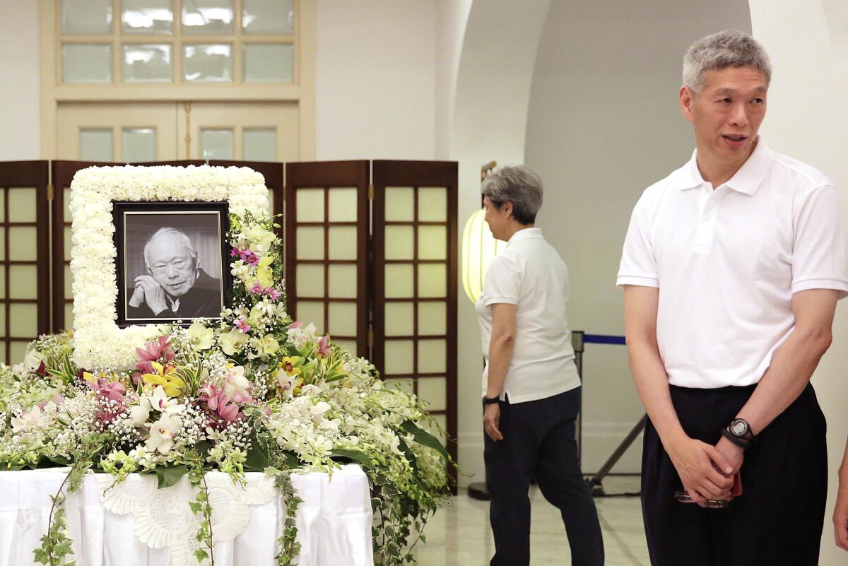 Two men in white shirts stand near a man's framed portrait with funeral flowers.