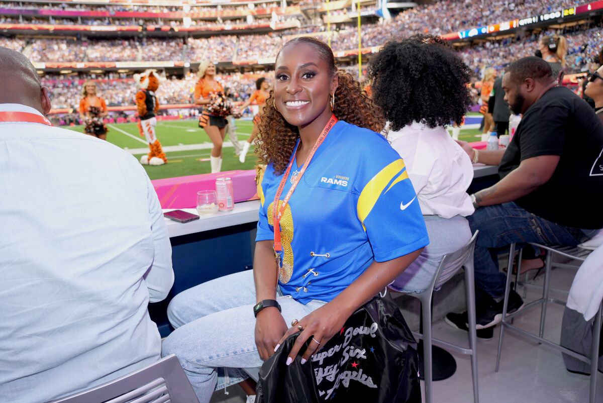 A smiling woman in a blue shirt sits at field level at a football game.
