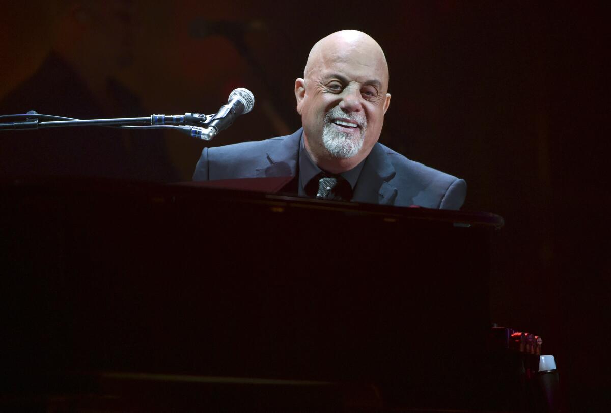 Billy Joel in a suit sitting behind a piano and smiling with a microphone near his face