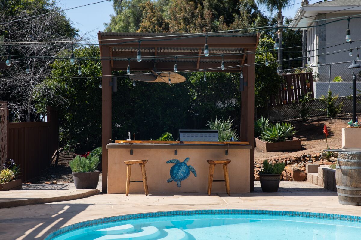 A remodeled outdoor kitchen near a pool has a roof with a ceiling fan, stools, a wide counter and new grill.