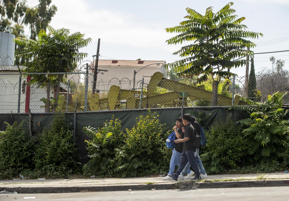 Oil pump jacks behind a razor-wire- topped chain-link fence with youths walking past on a sidewalk  