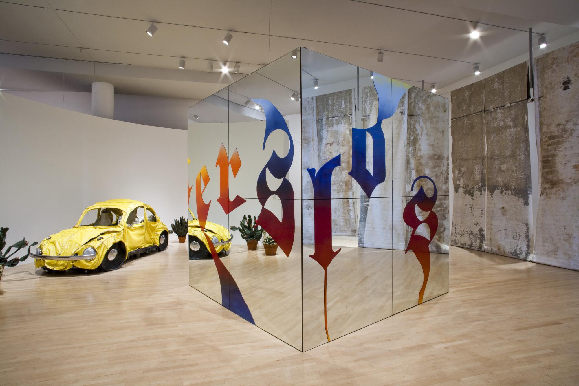 A view of a museum gallery shows "Migrant Dubs," a mirrored cube emblazoned with an Old English font.