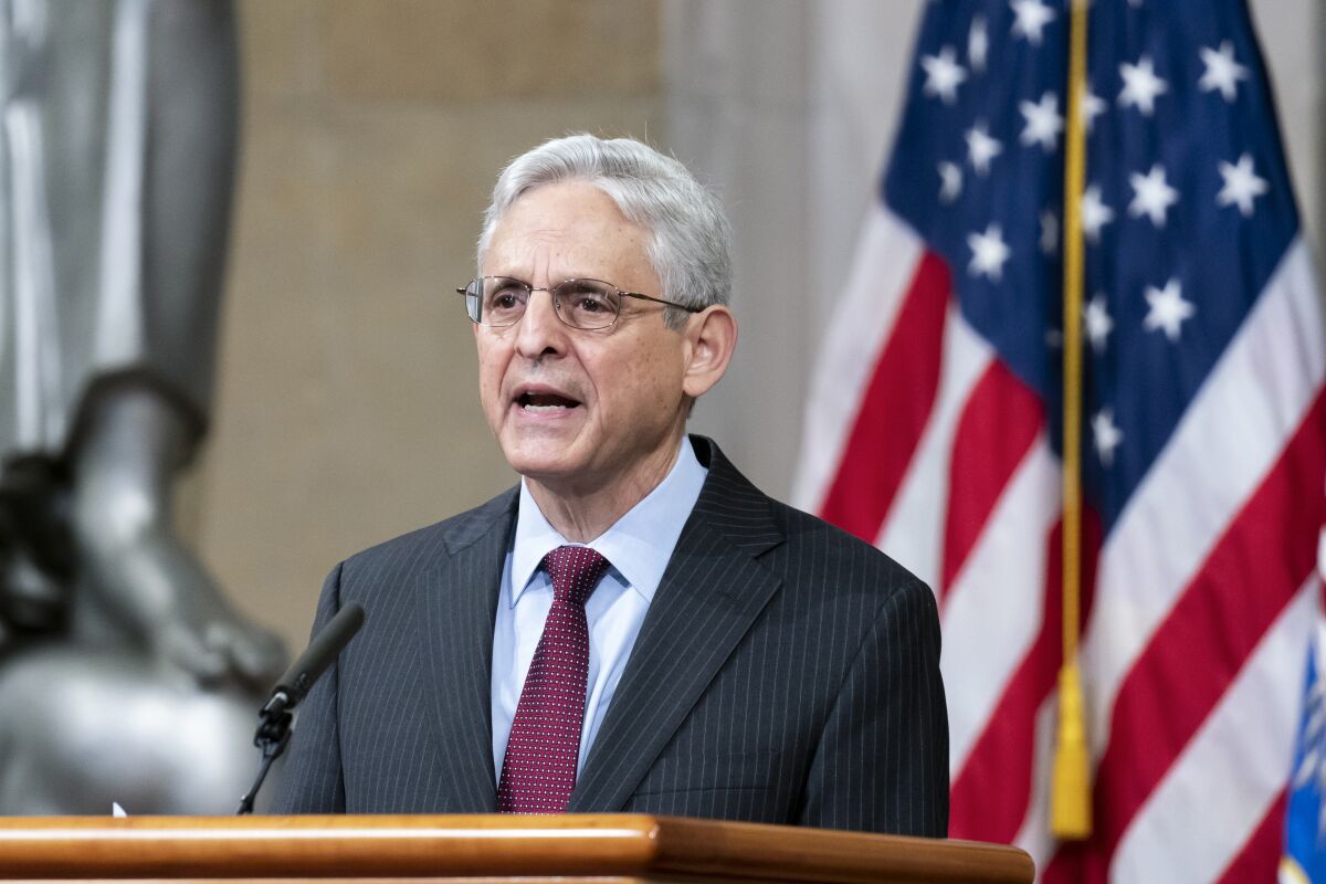 A man in glasses and a suit speaks in front of an American flag.