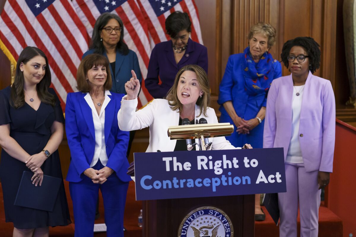Six women look on as another woman speaks at a podium that says "The Right to Contraception Act."