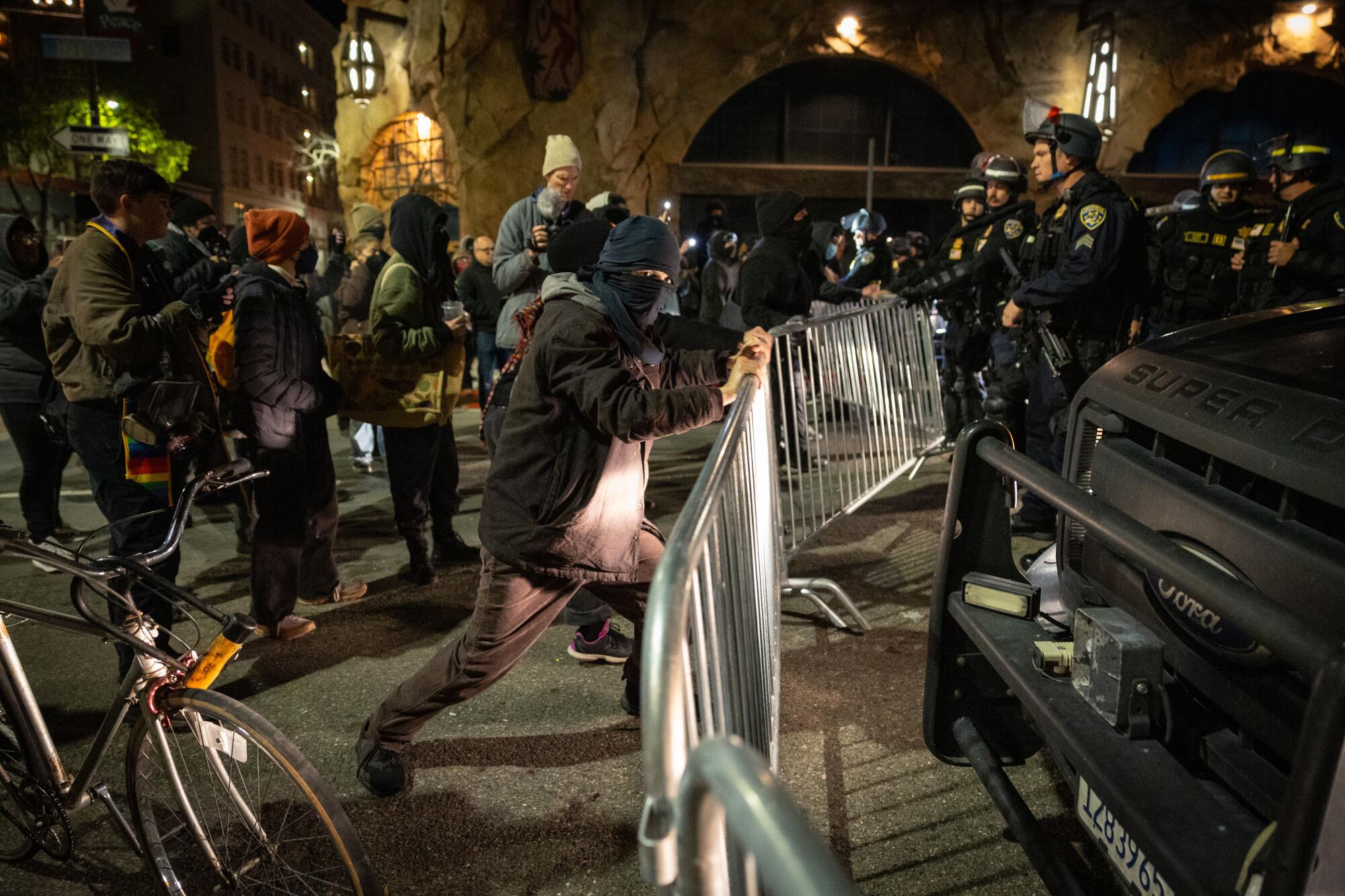 A masked man among a group of protesters wrestles with a metal crowd-control barrier as police look on