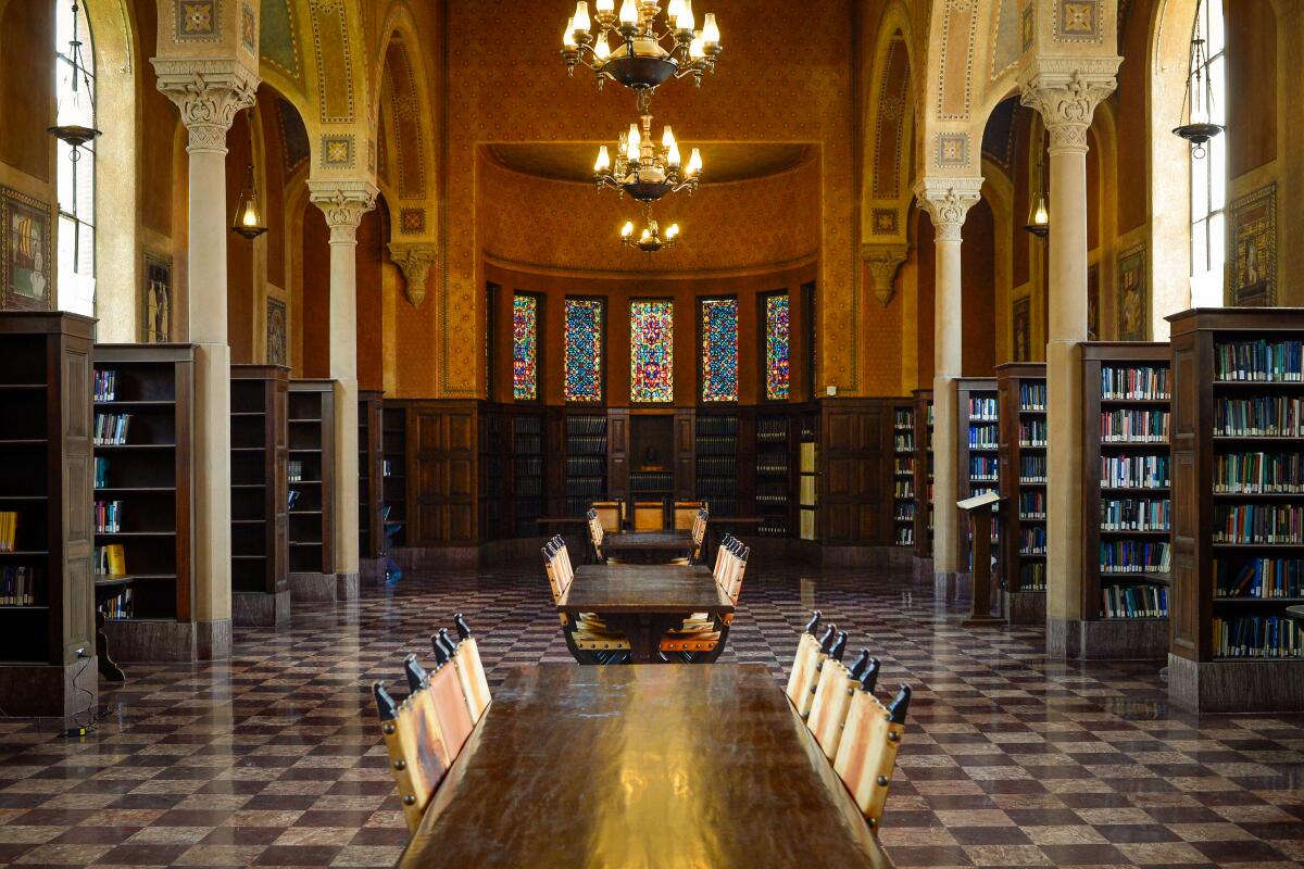An ornate room with checked tile floor, long tables, chandeliers and columned arches over bookshelves down the sides.