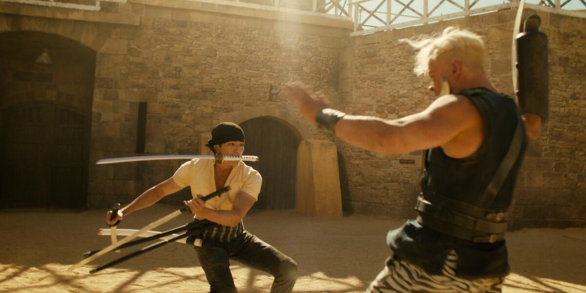 A man wearing a headwrap, white shirt and pants swings swords with his hands and mouth as a blond man in a vest falls.