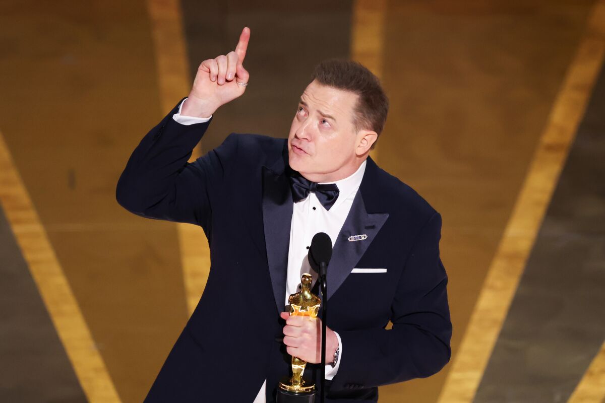A man in a suit holds an Oscar and points up