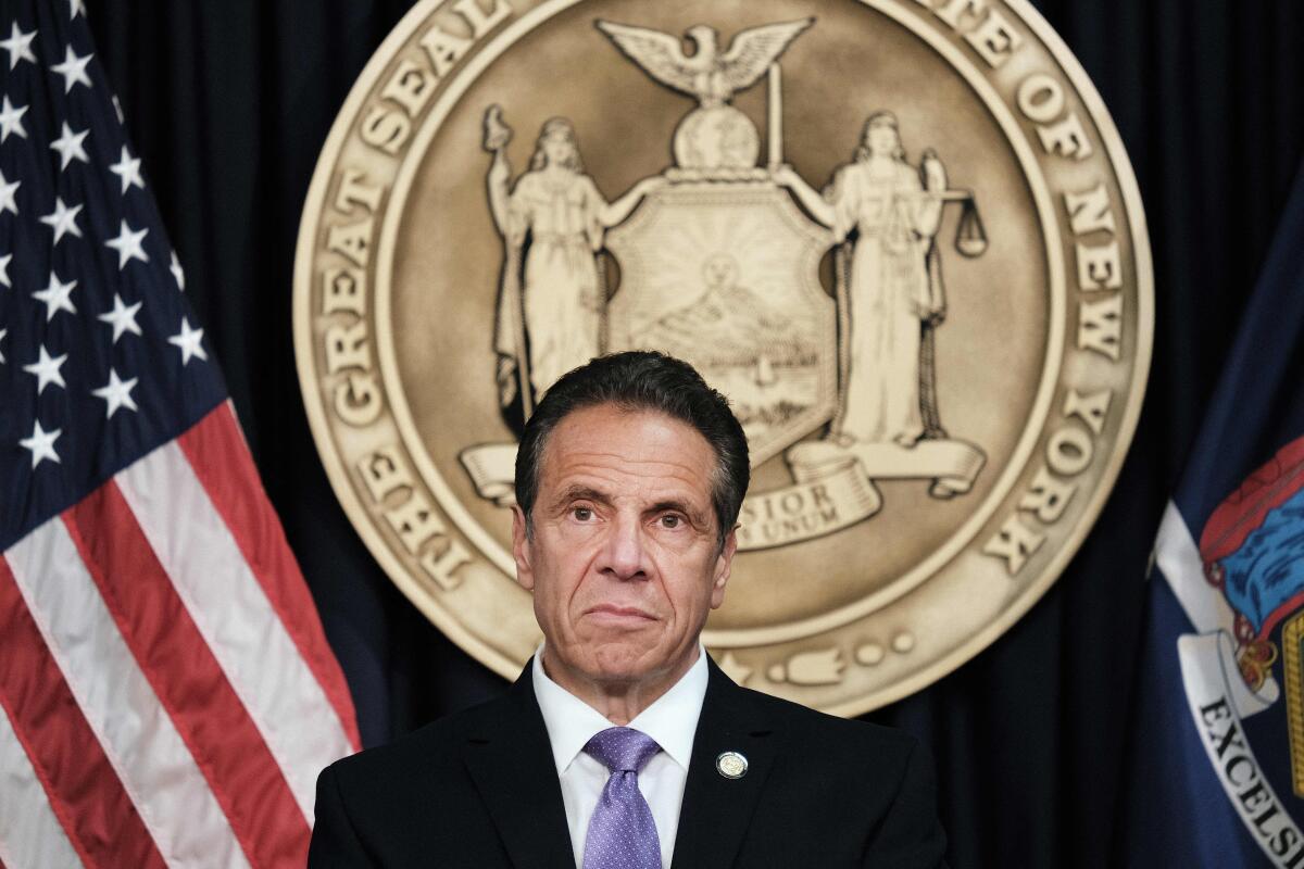 A closeup of an unsmiling Andrew Cuomo in front of the great seal of New York.