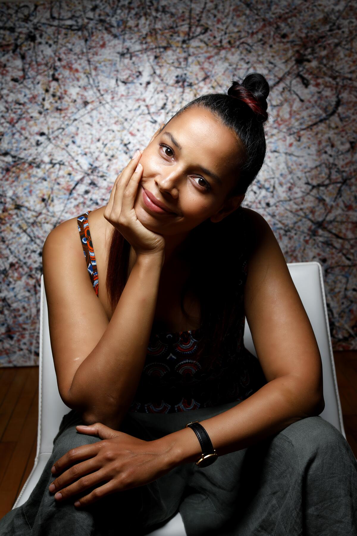 A woman, with her hair pulled back, sits in a white chair and leans her chin in her hand and gently smiles.