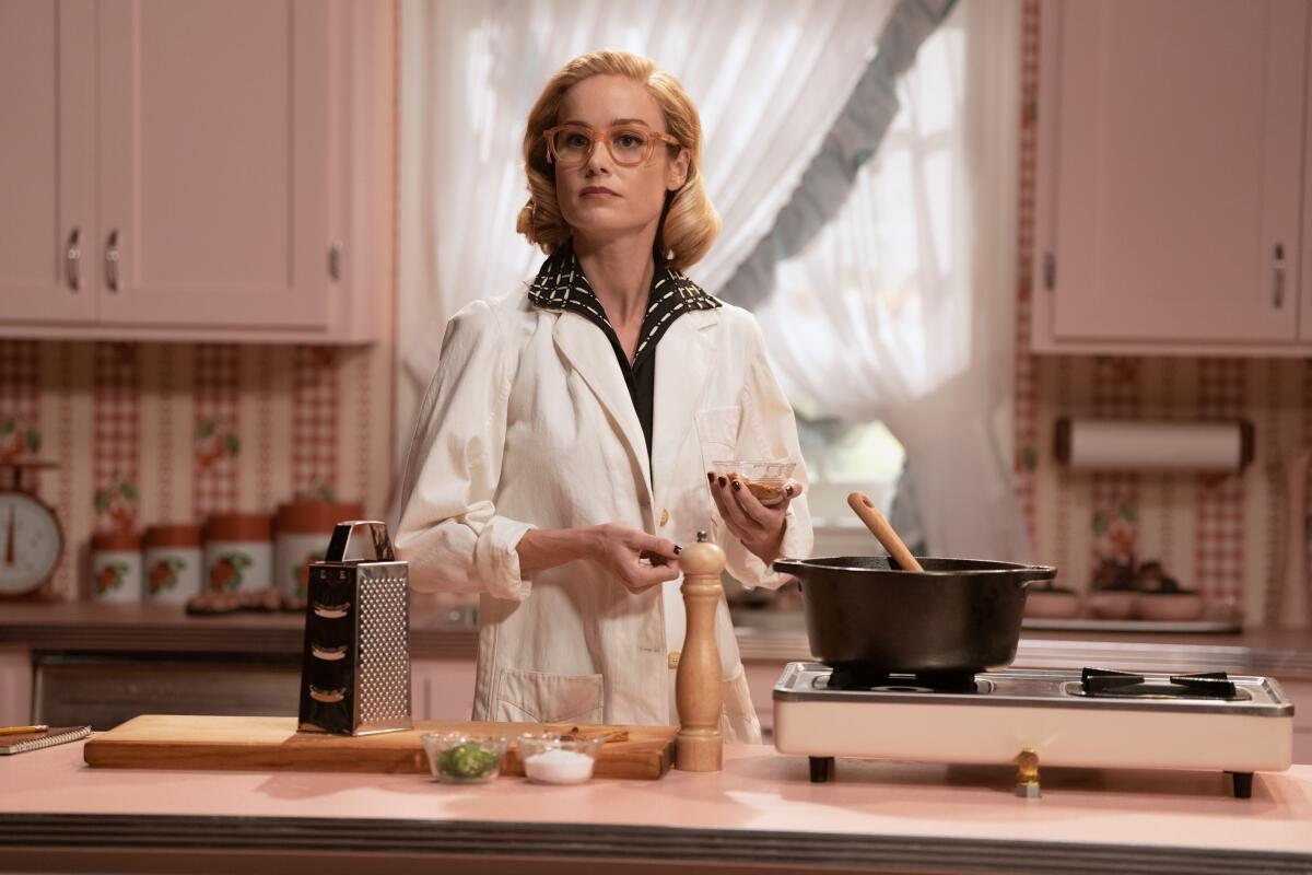 A woman in a white coat and glasses cooks in a pink kitchen