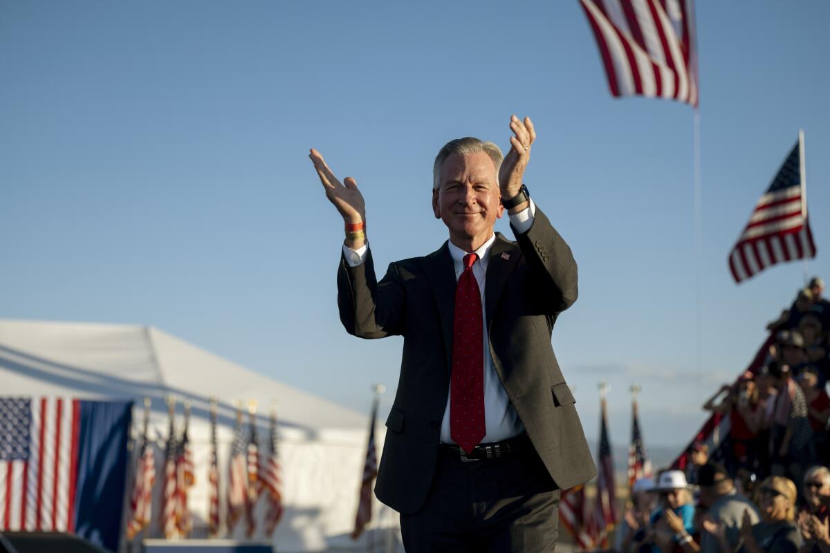 Sen. Tommy Tuberville raises his hands outdoors near a crowd and U.S. flags.