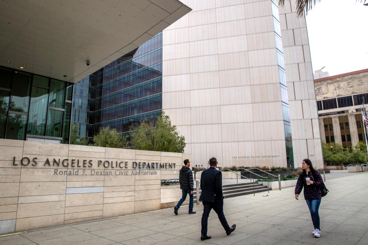 Three people walk by a building with the words "Los Angeles Police Department" on a wall.