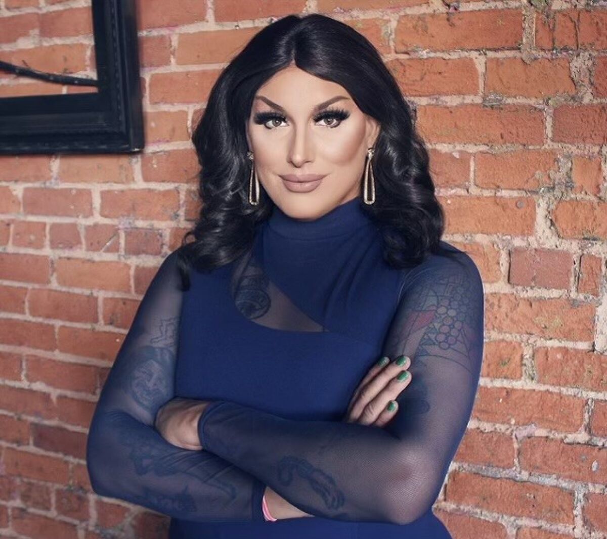 Maebe A. Girl, sporting dramatic makeup and earrings, poses for a photo with arms crossed, against a brick wall.