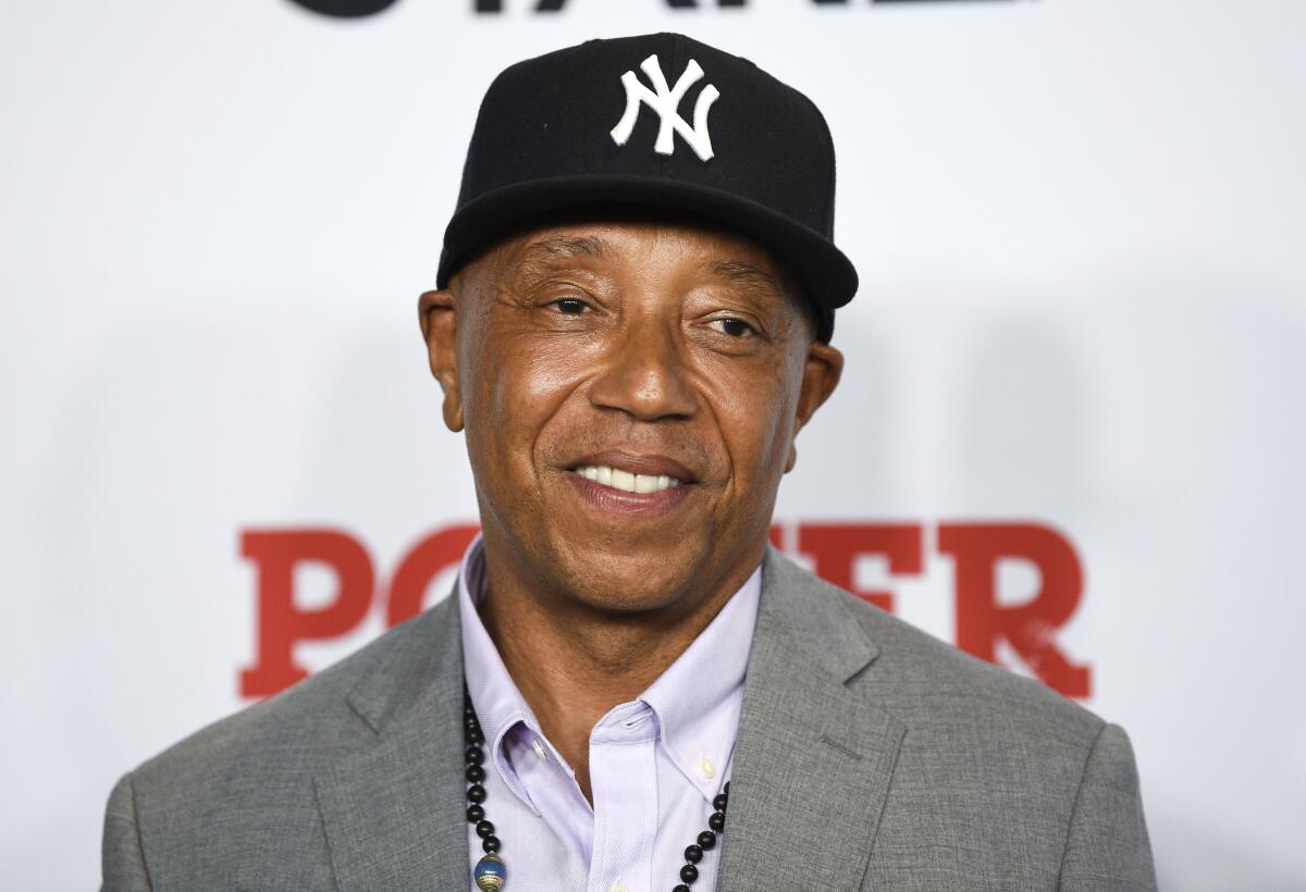 Russell Simmons wears a black baseball cap and gray blazer with a beaded necklace. 