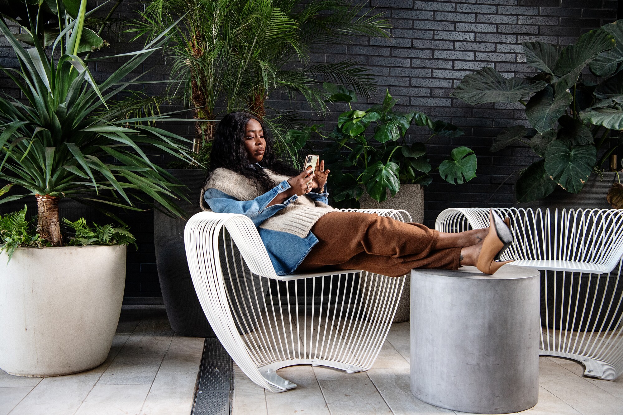 A woman sits on a white wire chair with her feet up on a cylindrical concrete table.