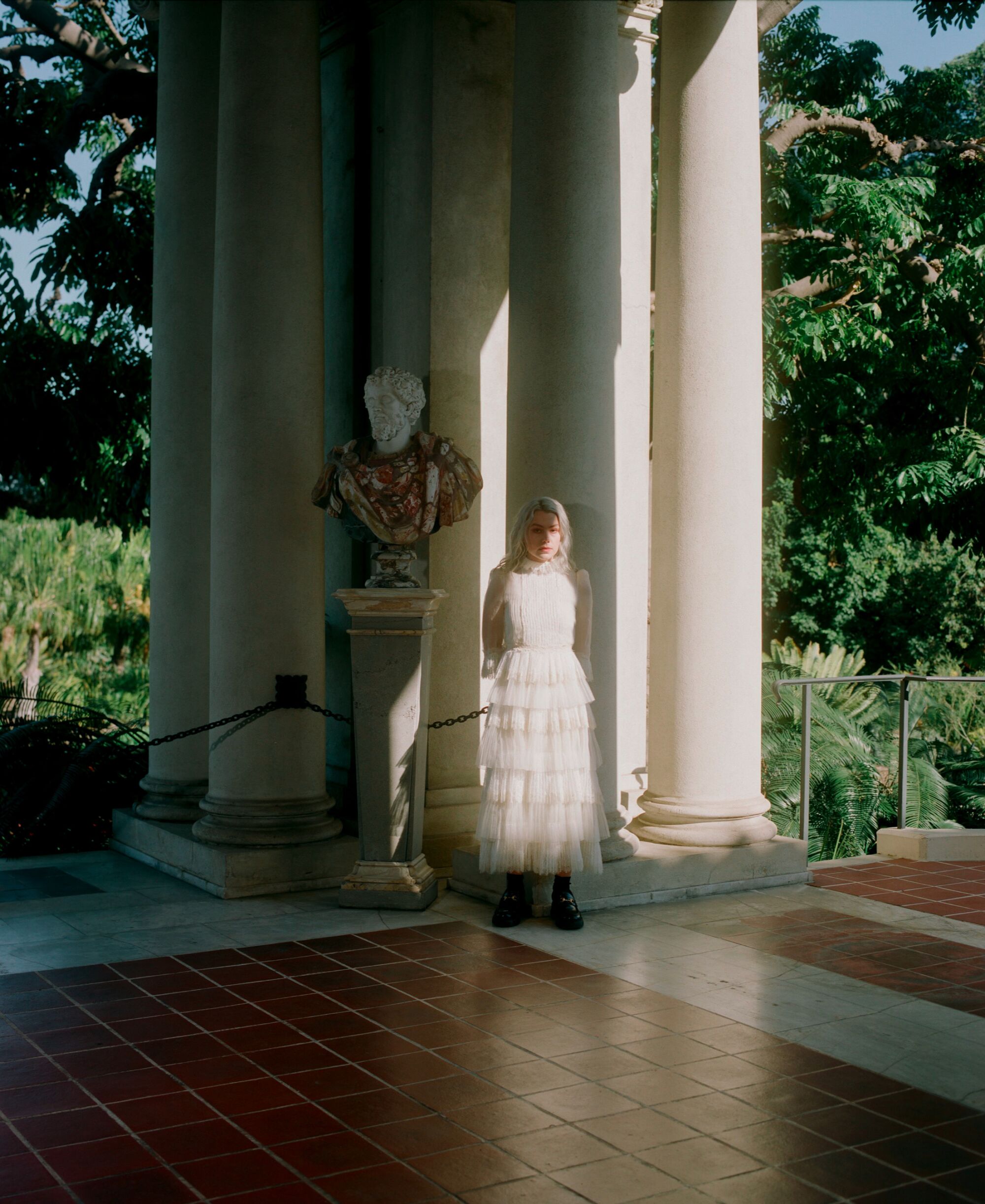 A woman in a white dress leans against a column next to a stone bust of a man.