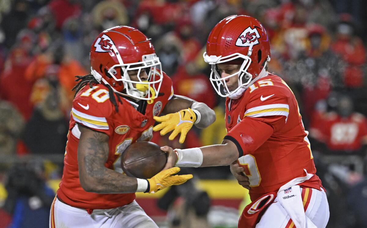 Two football players in red jerseys and helmets, one handing the ball off to the other