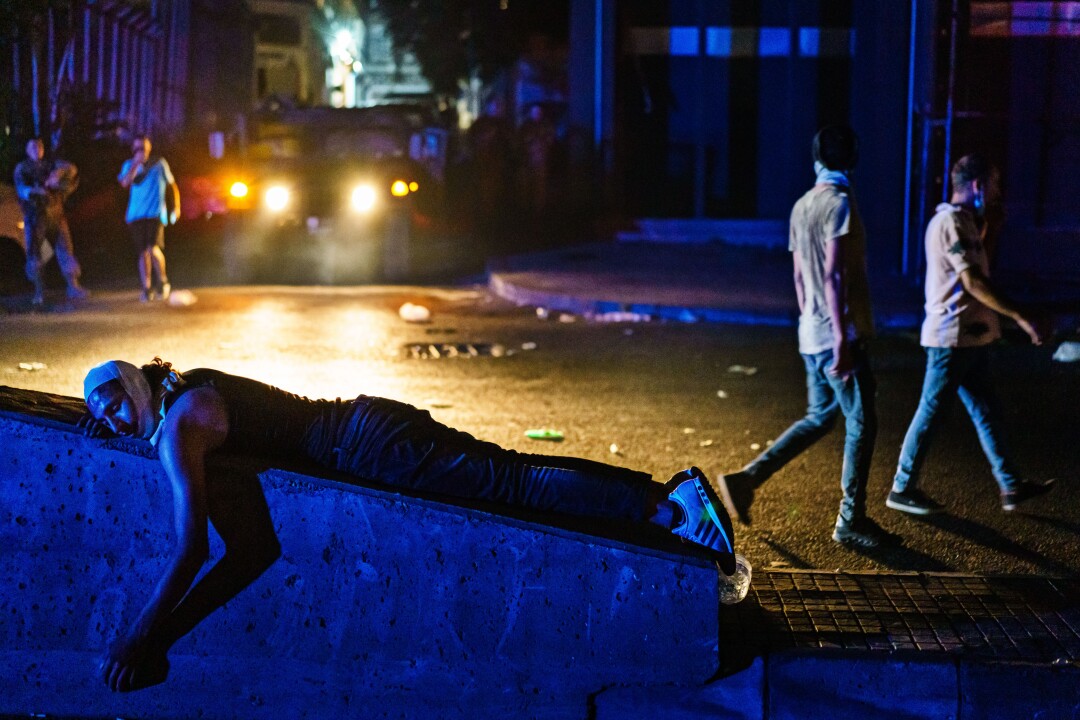 At night, a man lies on his stomach on a concrete block as people walk in the street behind him.