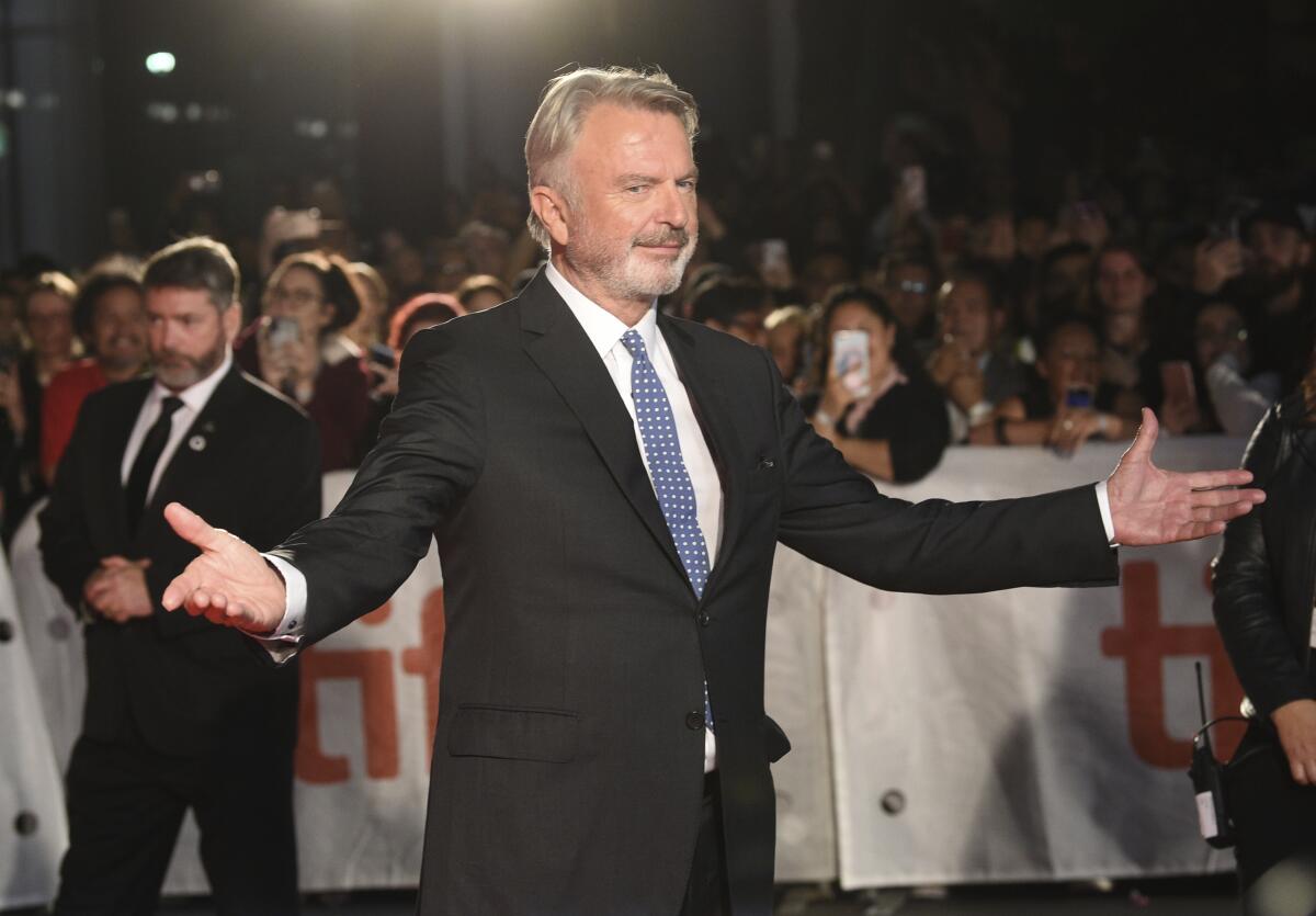 A man with gray hair and a beard wearing a black suit and posing with his arms outstretched in front of a barricaded crowd