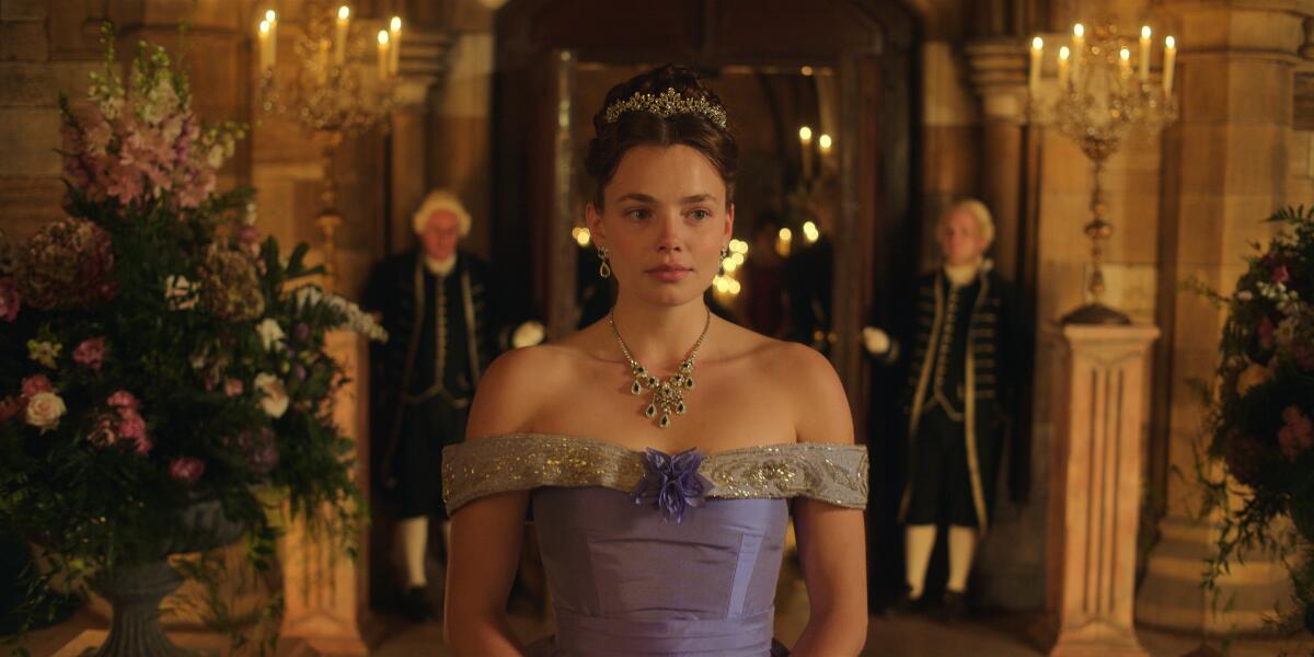 Nan in a bare-shouldered purple dress, tiara and jeweled necklace walks down a hallway.