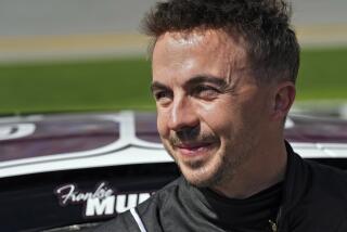 Frankie Muniz dressed in black smiles in the sun, standing in front of his race car at the Daytona Speedway