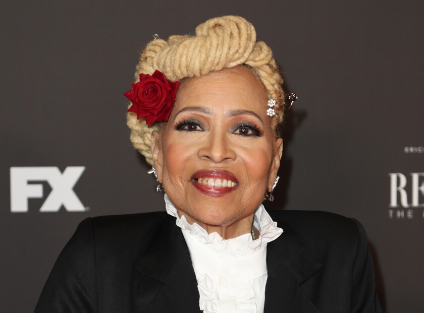 A woman with blond hair wrapped into a bun with a red rose in it posing in a black suit and ruffled white shirt