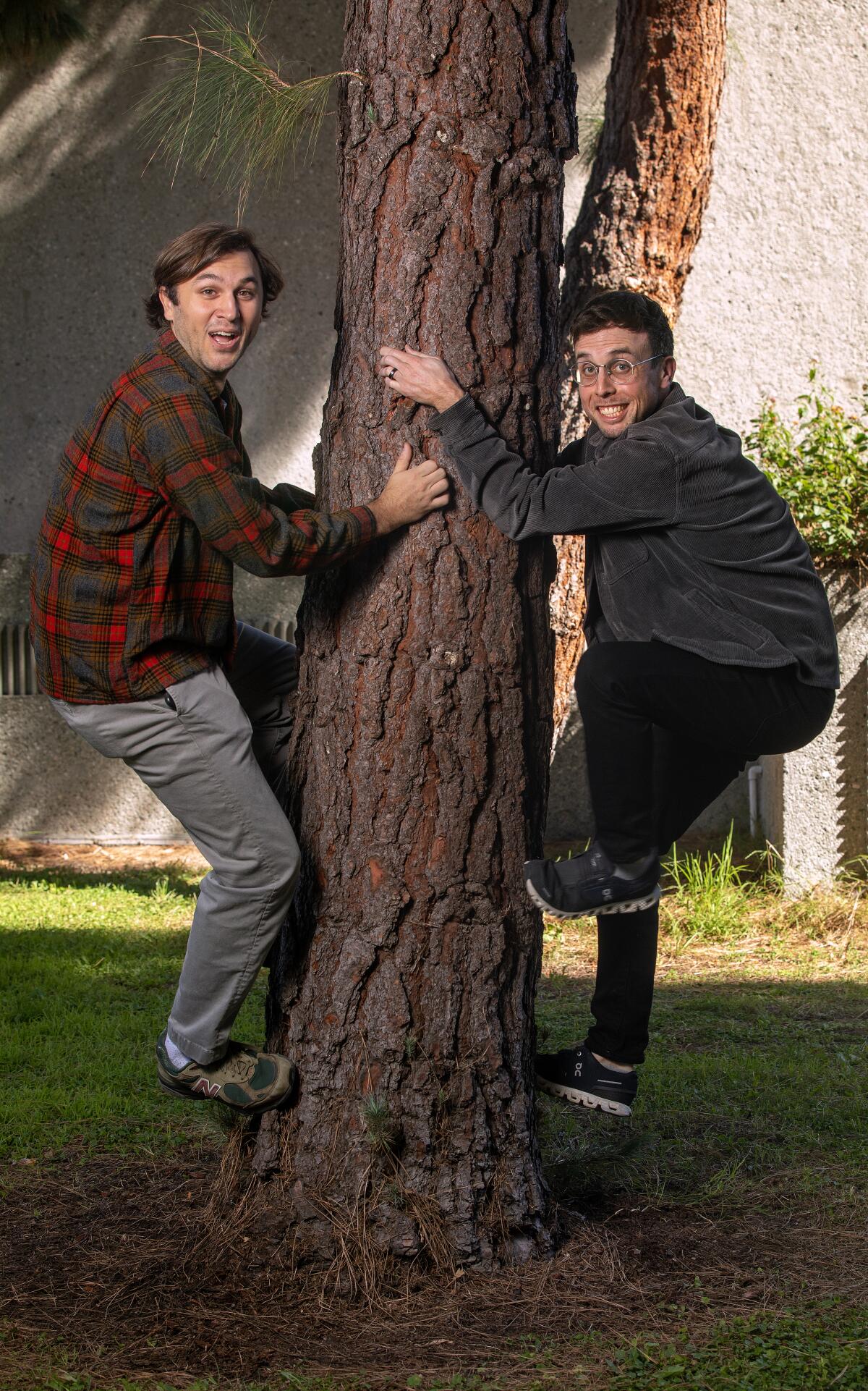A man in a flannel shirt and one in a dark gray/green shirt make faces as they pretend to climb a tree in Barnsdall Art Park.