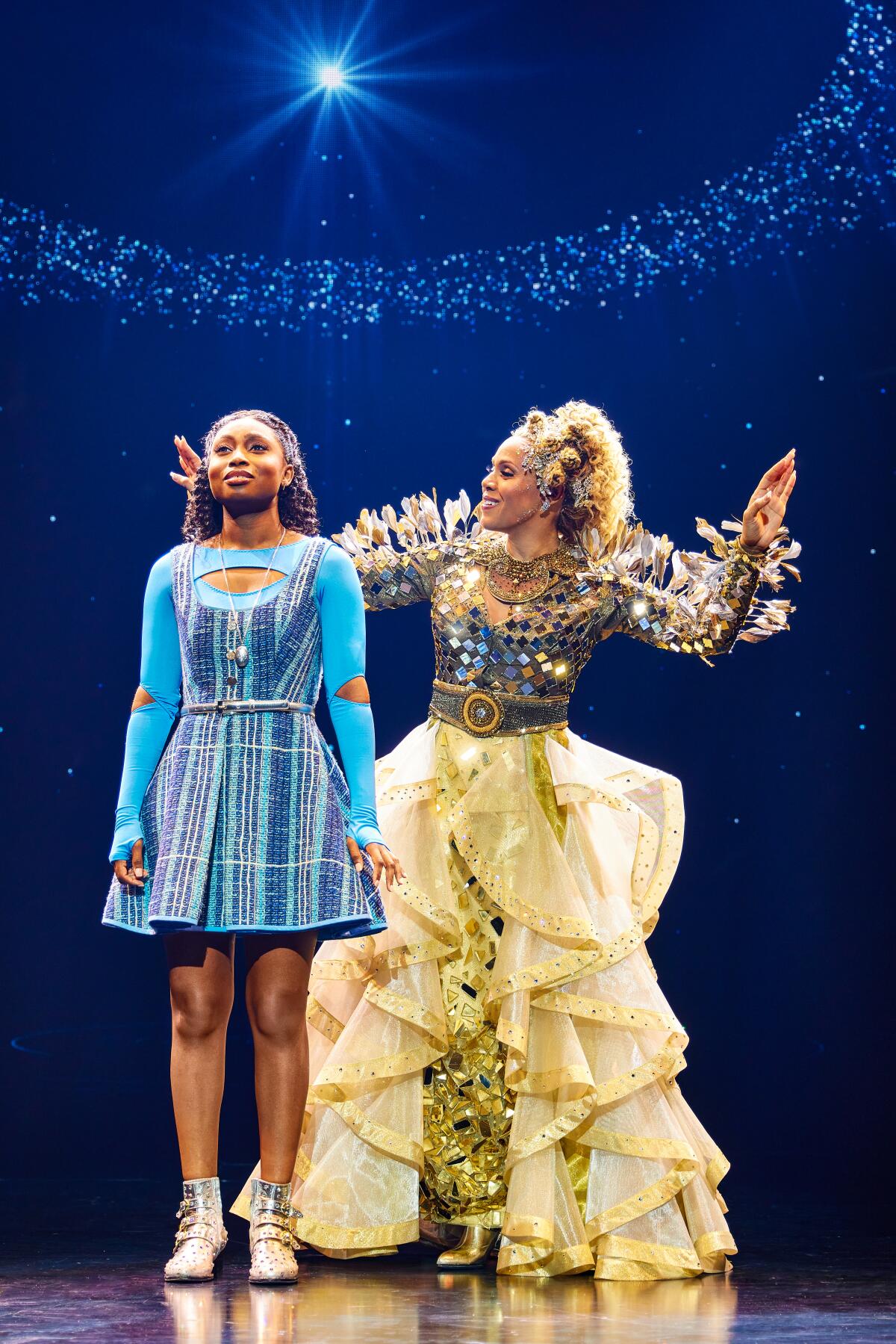 A woman in an elaborate costume sings to a girl in a blue dress on stage