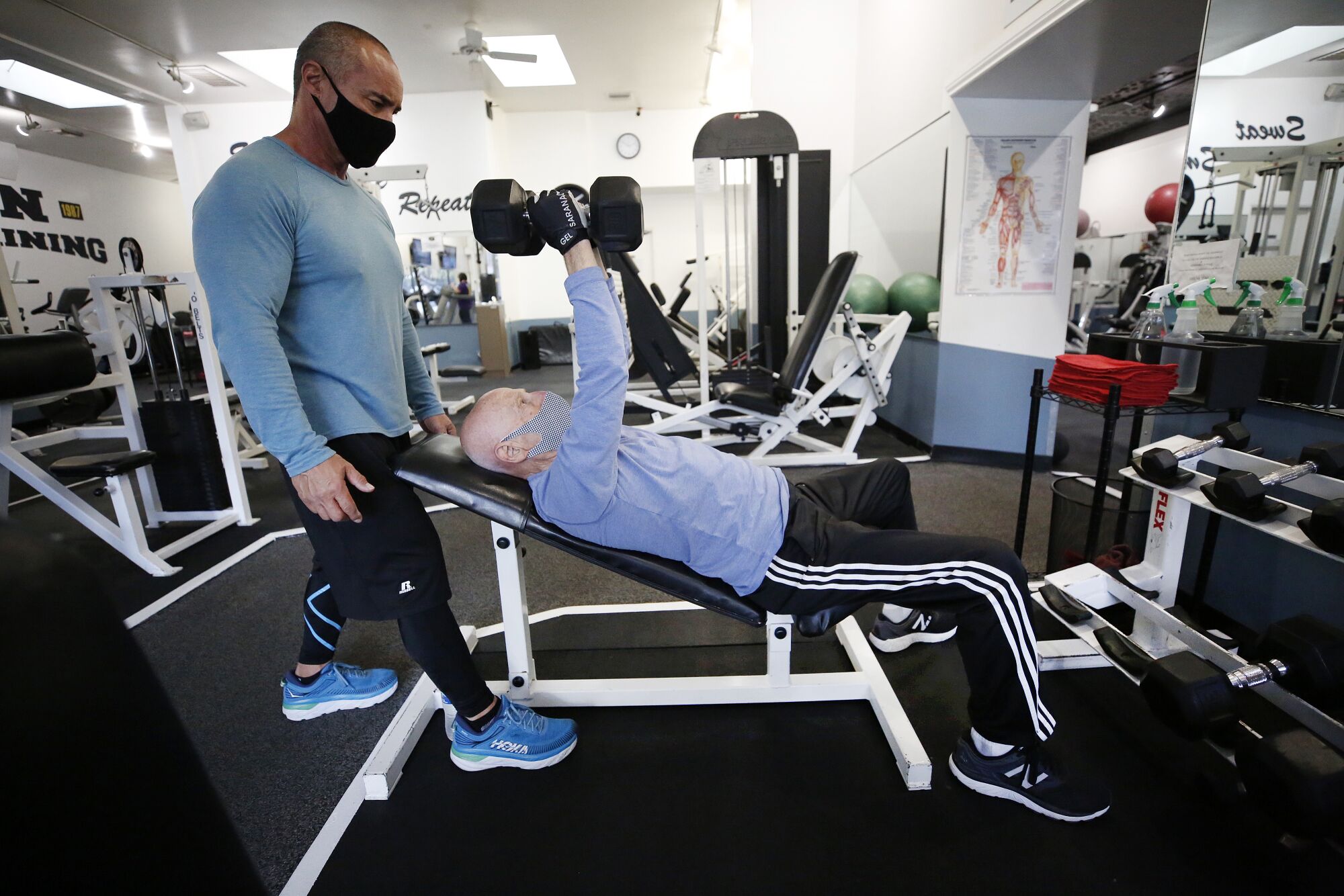 A masked man on a bench lifts dumbbells as another man, left, watches 