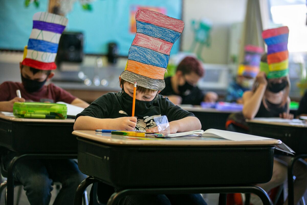 Students sit at desks completing schoolwork while wearing masks and Dr. Seuss hats.