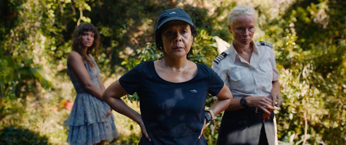 An unhappy-looking woman stands, hands on her hips, with two women behind her among foliage.