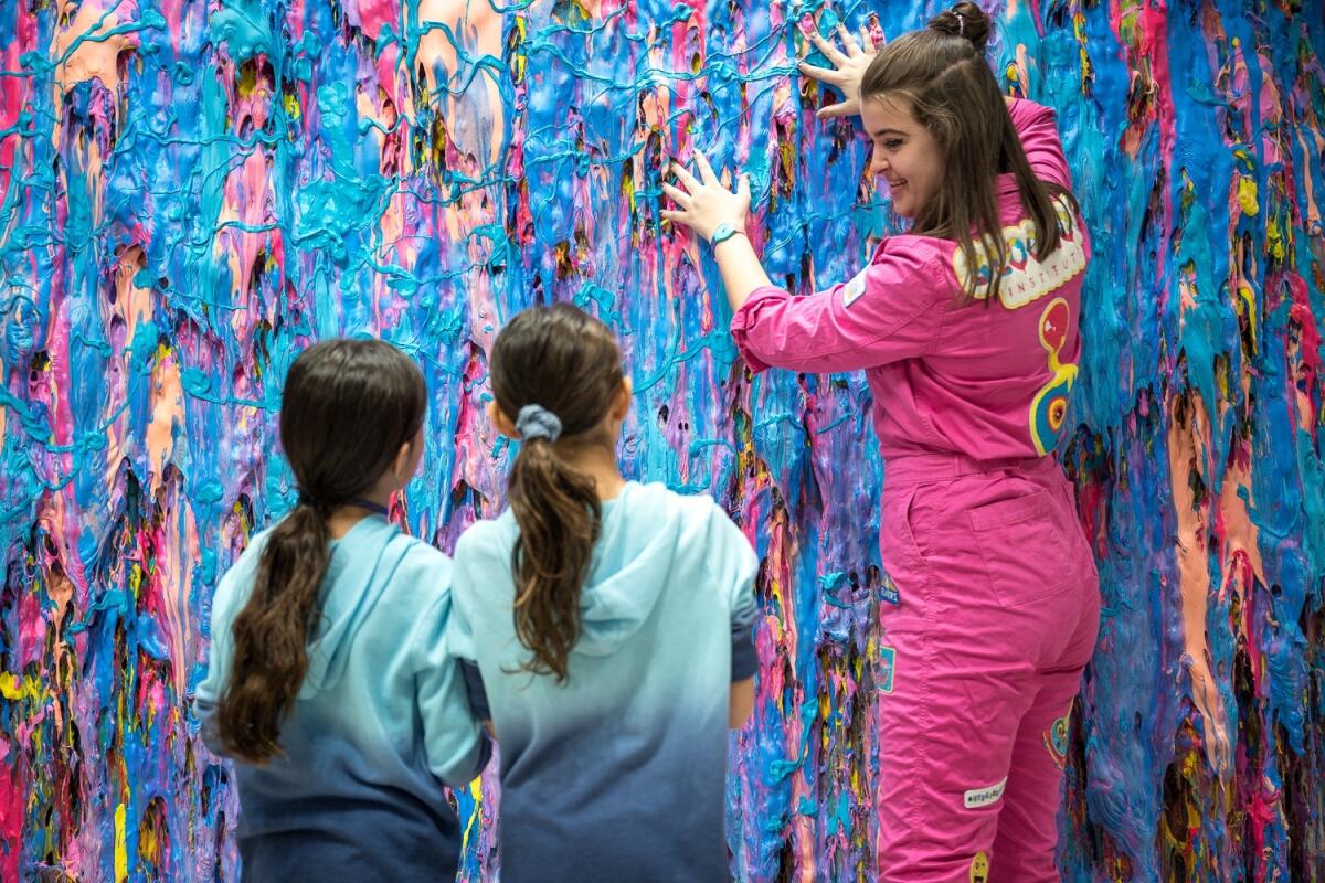 Three girls look at and touch a wall covered in slime.