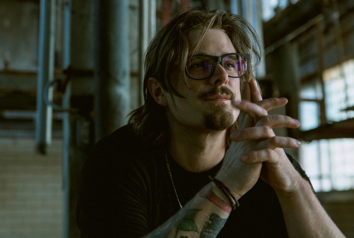 A man with long shaggy hair, a black t-shirt and glasses sits, his hands folded in front of him.